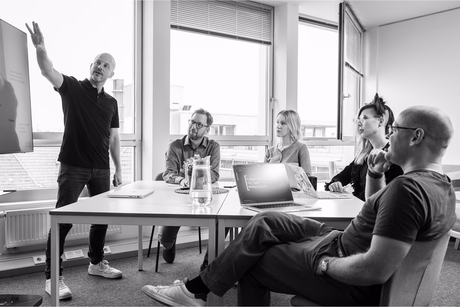 Das intcube-Team bei einem Meeting in einem hellen Büro. Eine Person steht neben einem Bildschirm mit einer Präsentation zu Resilienz und digitaler Souveränität, die anderen hören zu und lachen gemeinsam.