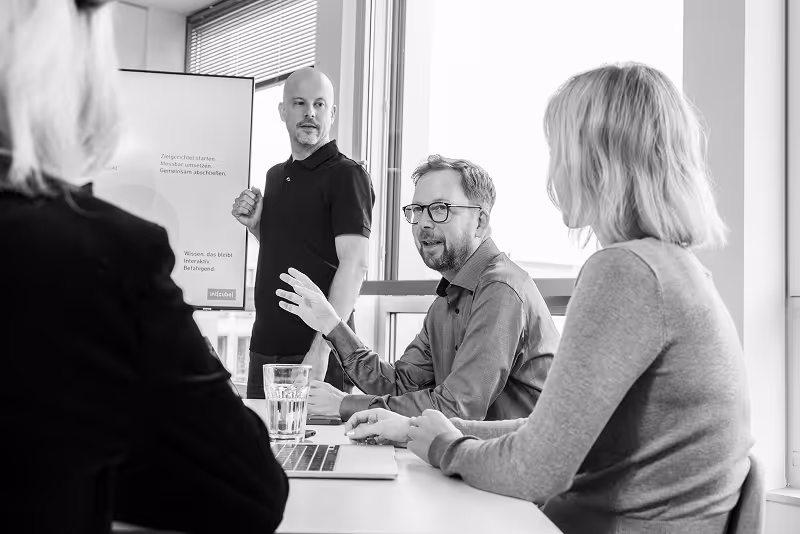 Teammeeting mit drei Personen an einem Tisch, im Hintergrund ein Whiteboard.
