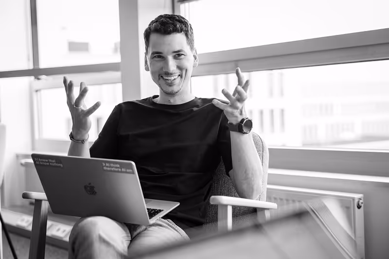 Mitarbeiter gestikuliert lächelnd in einem Lounge-Bereich mit Laptop.